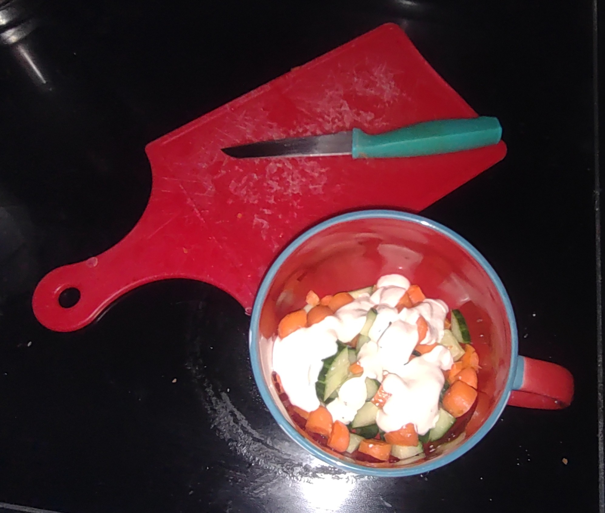 picture of diced carrots and cucumbers in a large mug slathered in ranch dressing, in the background is a red cutting board with a knife on it