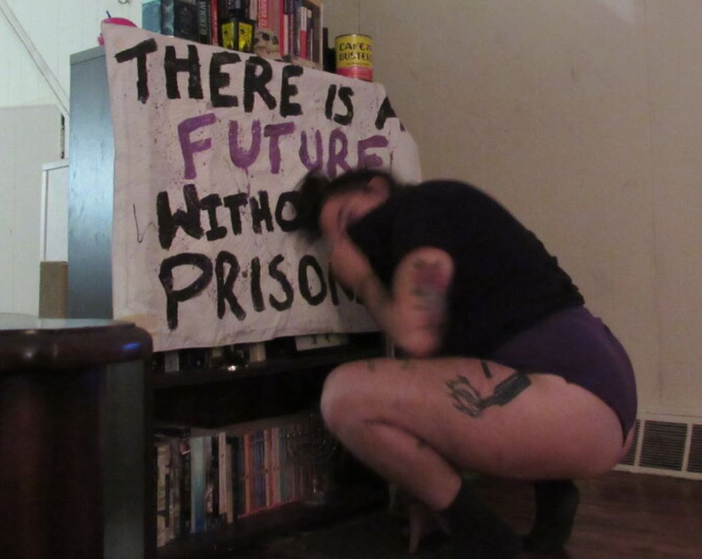 a photo of me, white curvy brunette tattooed adult, squatting in front of a banner that says: THERE IS A FUTURE WITHOUT PRISONS